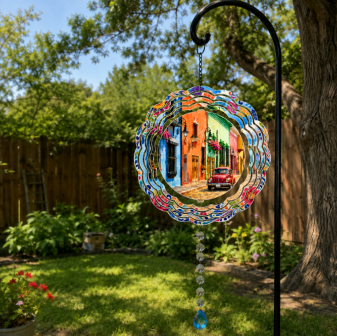 A circular stained-glass style garden suncatcher hanging from a black metal garden stake in a backyard garden. The suncatcher features a colorful, swirling border and a central printed image of a vibrant street scene with colorful houses and a red car.
