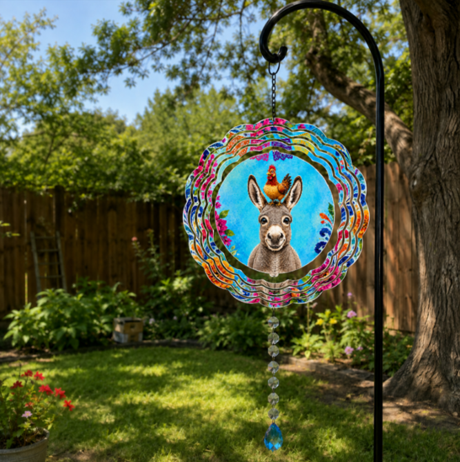 A circular metal garden suncatcher hanging from a black garden stake in a backyard garden. The suncatcher features a colorful, wavy border with pink, blue, and orange patterns, a central illustration of a donkey on a blue background with a small rooster on its head, and a dangling beaded tassel with a blue crystal at the bottom.