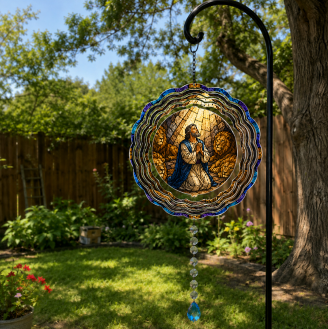 A stained glass suncatcher featuring a praying man surrounded by lions, hanging from a black metal garden stake in a sunny backyard garden.