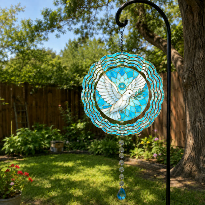 A circular stained-glass-style garden suncatcher hanging from a black metal garden stake in a backyard. The design features a white dove with outstretched wings over a bright turquoise and blue floral pattern, with a dangling clear bead chain ending in a blue teardrop.