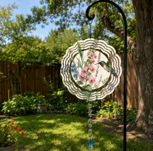 A circular metal garden suncatcher with a scalloped edge, featuring a printed design of a green hummingbird, pink foxglove flowers, and blue lavender, hanging from a black metal garden stand in a sunny backyard garden.