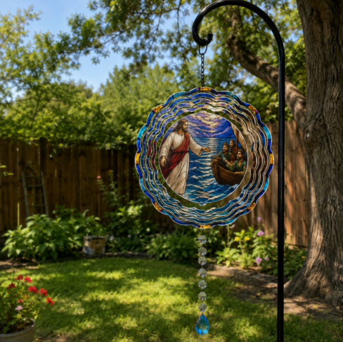 A circular stained-glass style garden suncatcher hanging from a black metal garden stake in a backyard garden. The suncatcher features a colorful depiction of Jesus reaching out of a boat with blue, gold, and red glass accents, and a dangling clear glass bead teardrop at the bottom.