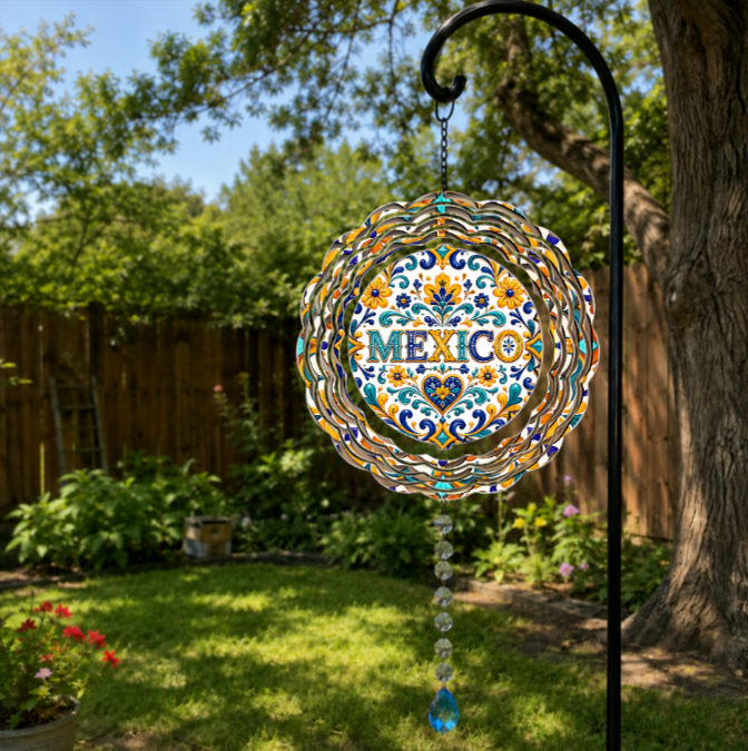 A circular glass Mexican-themed garden suncatcher hanging from a black metal garden stake in a backyard. The suncatcher features a colorful Mexican folk art pattern with the word 'MEXICO' in the center and a dangling blue glass bead tassel.