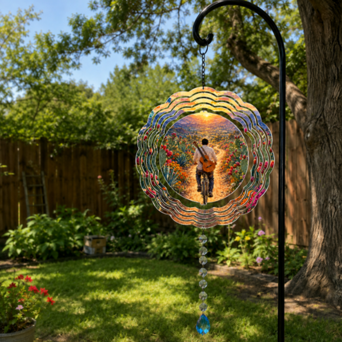 A circular glass suncatcher with a colorful sunset and a cyclist design, hanging from a black metal garden stake in a lush green backyard.