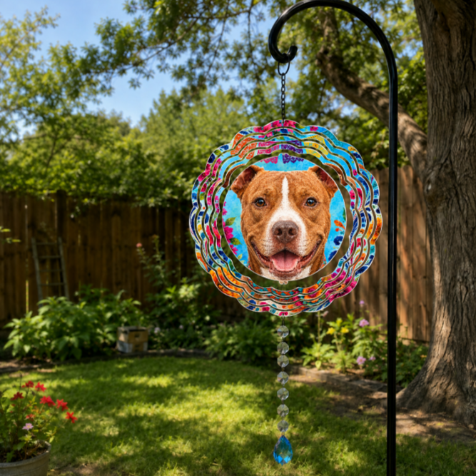A circular metal garden suncatcher with a colorful marbled border and a portrait of a brown and white dog in the center, hanging from a black metal garden stake in a sunny backyard garden.